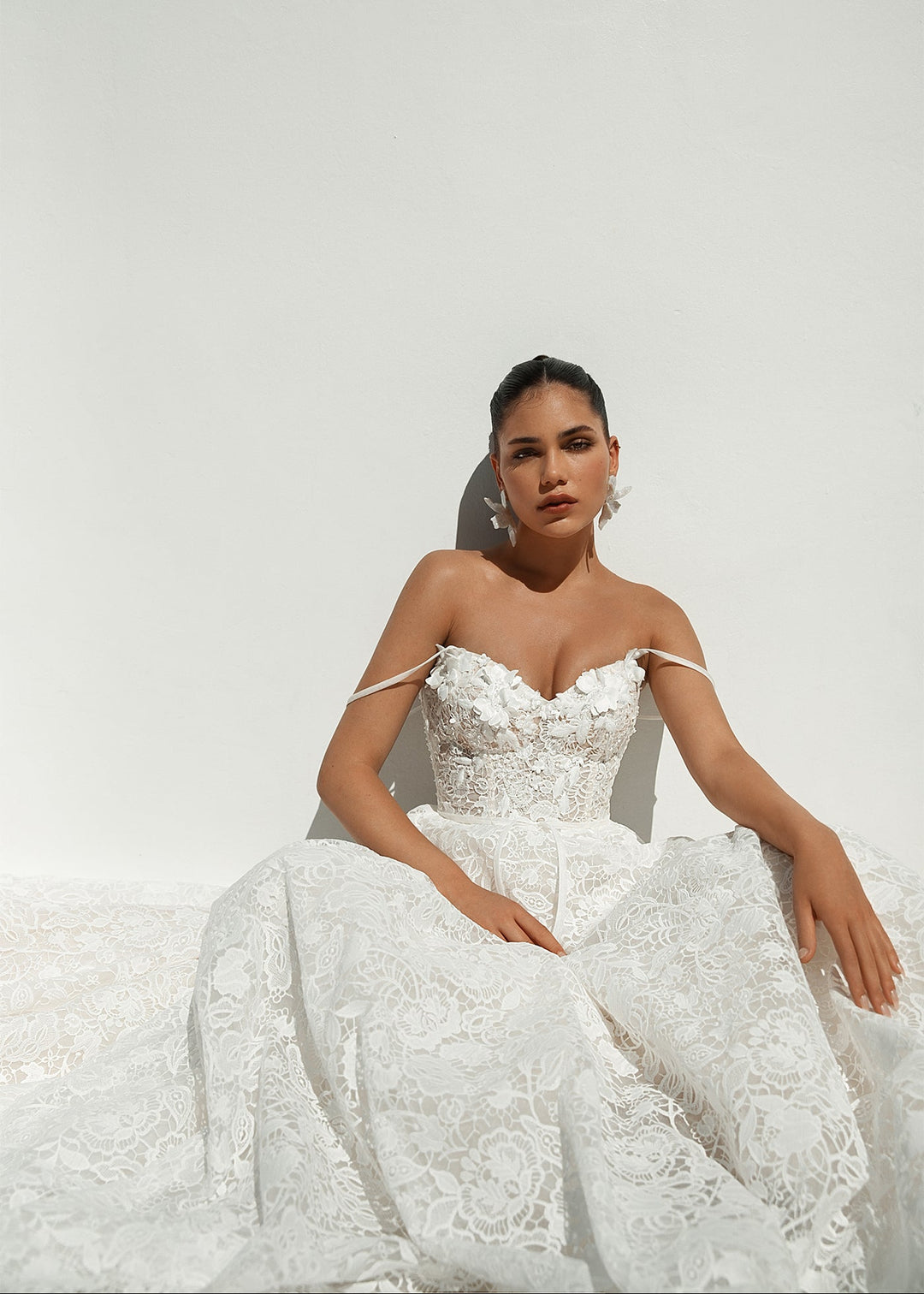 Aeris gown with all over lace and 3D flowers, shown on a petite model in a front view sitting against a rendered wall.