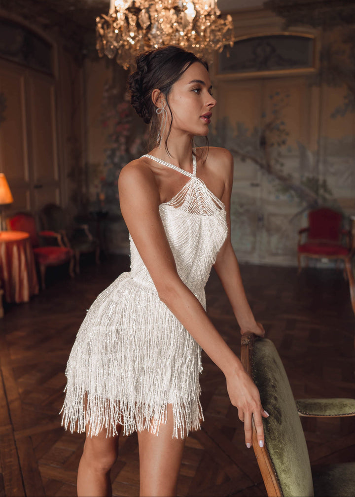 Woman wearing a sequin fringe mini dress  at her wedding reception leaning against a chair in an ornate room.