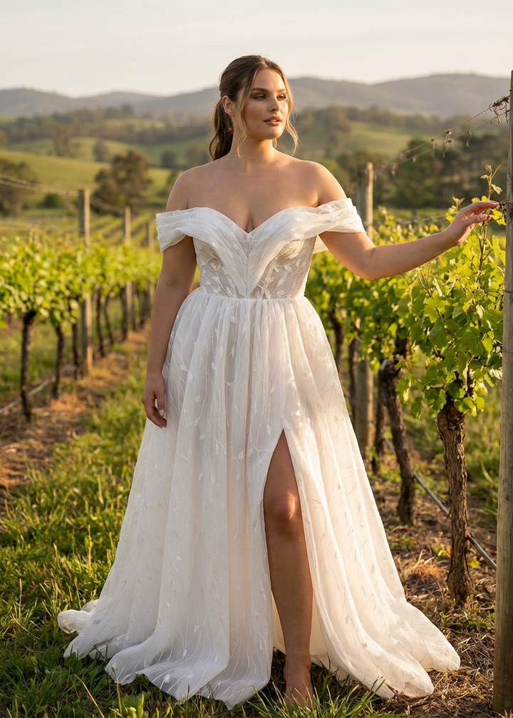 Woman in an off-the-shoulder pleated botanical tulle A-line wedding dress with a leg slit standing in a vineyard.