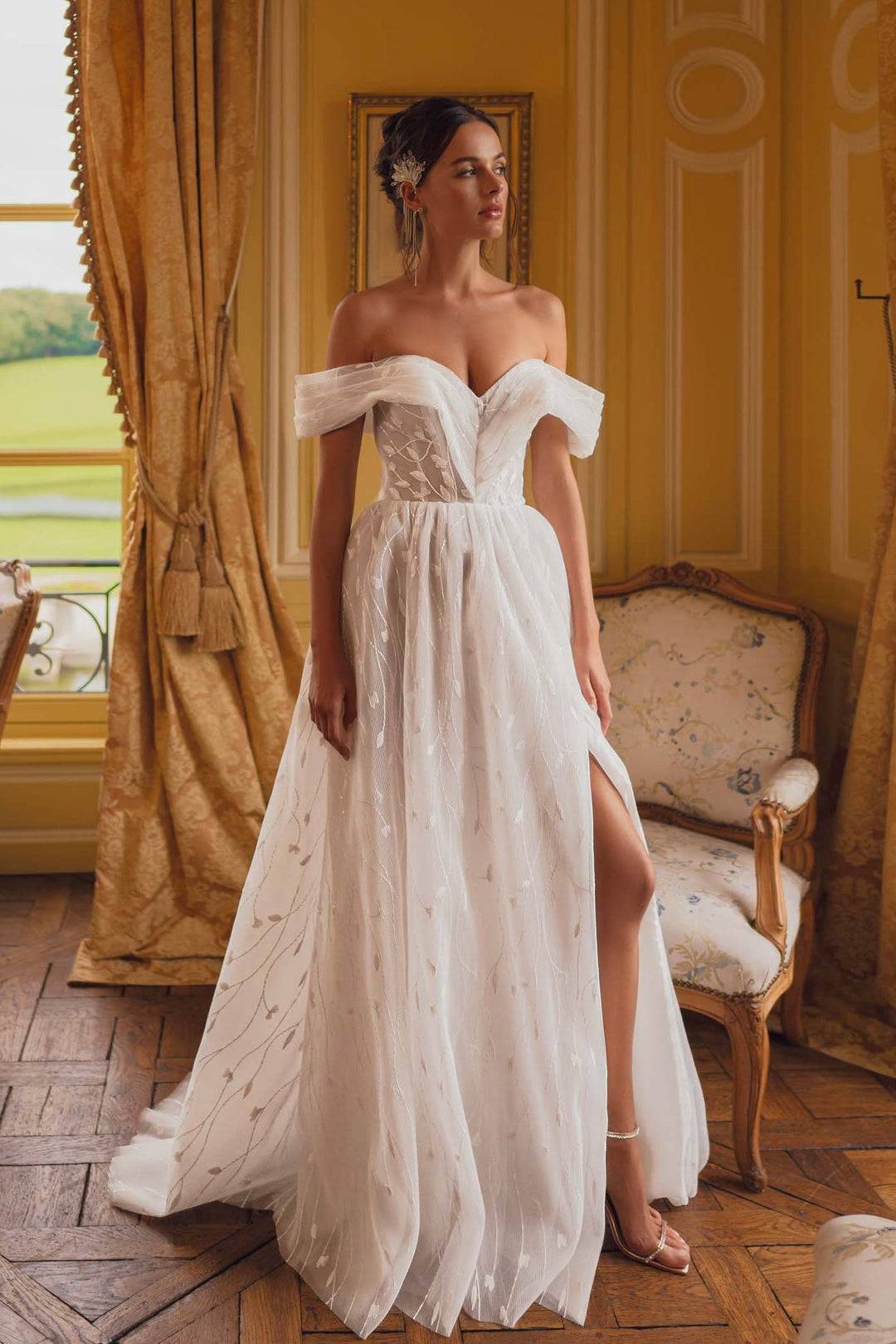 A woman standing in a room with classic decor, wearing an off-the-shoulder, floral embroidered, A-line wedding dress with a thigh-high split on one side.