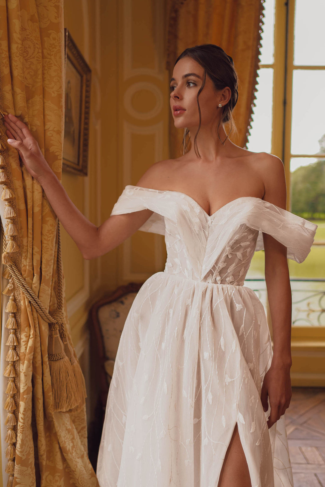 Woman in an off-the-shoulder pleated botanical tulle A-line wedding dress with a leg slit standing in a room.