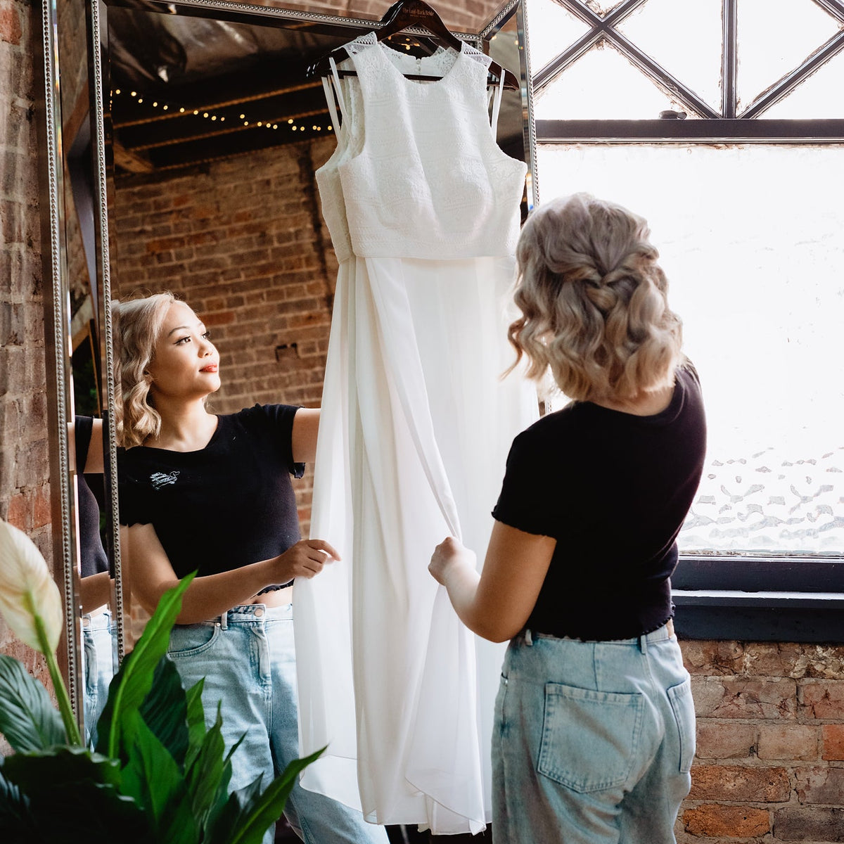 A woman in a black t-shirt and light blue jeans stands in front of a large mirror in a loft-style room with exposed brick walls, holding out a sleeveless white wedding dress hanging on a hanger.