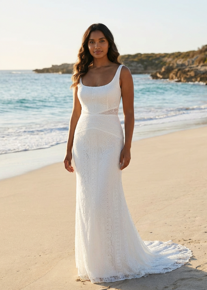 Woman in a white column lace dress standing on a beach with ocean and sky in the background.