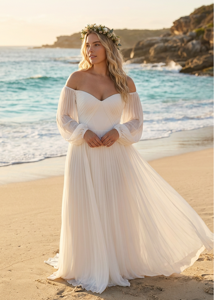 Woman in a pleated chiffon wedding dress standing on a beach with ocean waves in the background.