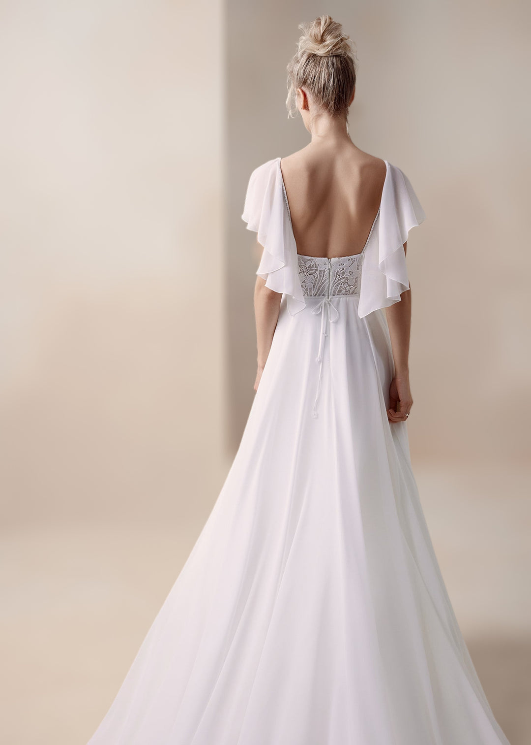 Woman in a chiffon a-line wedding dress with a modern lace bodice, open back and flutter sleeves, standing in front of a neutral background.