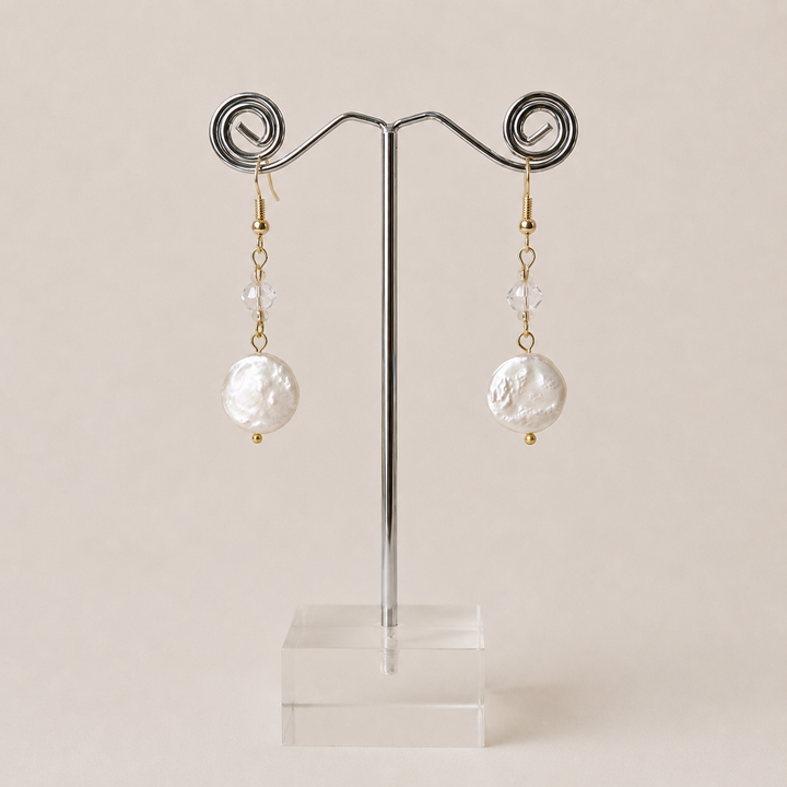 A pair of dangling earrings displayed on a metal jewelry stand against a warm beige background. Each earring has a gold hook, a small clear faceted bead, a gold spacer bead, and a large irregular coin-shaped freshwater pearl.