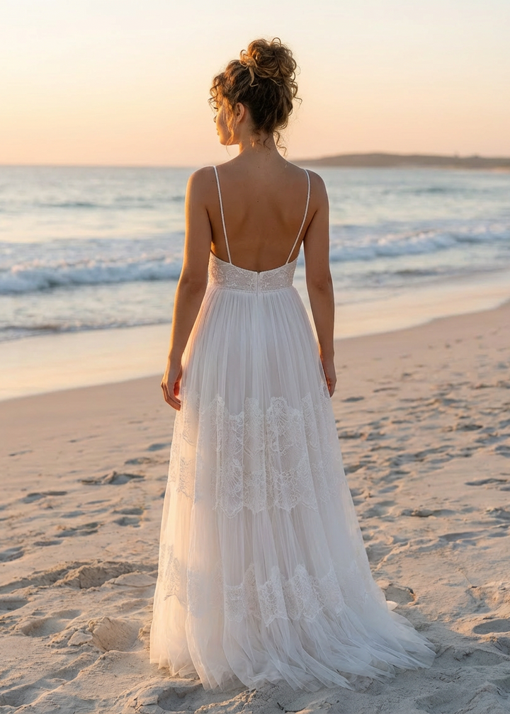 Woman in an a-line spaghetti strap lace and tulle wedding dress with a low open back standing on a beach at sunset.