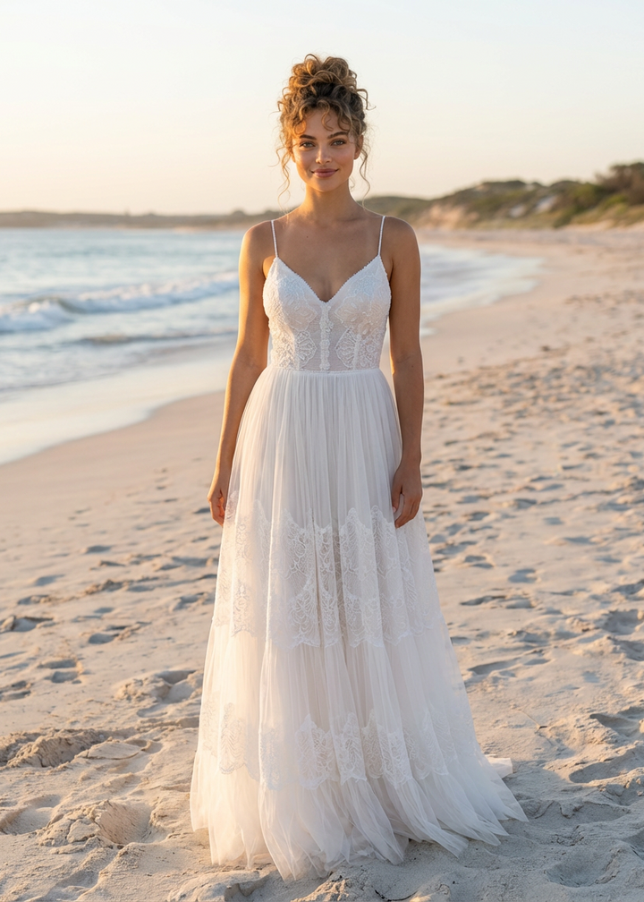 Woman in an a-line spaghetti strap lace and tulle wedding dress standing on a beach at sunset.