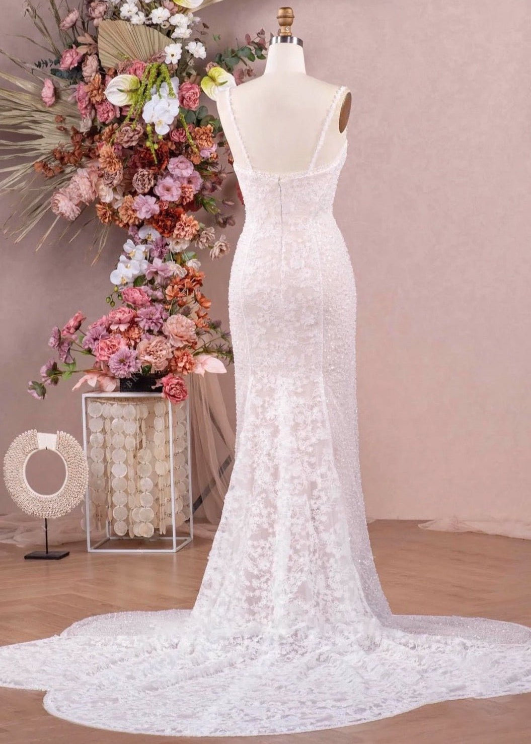 A fitted wedding dress with spaghetti straps, beading, lace and a flower cut-out train on a mannequin in front of a studio setting.