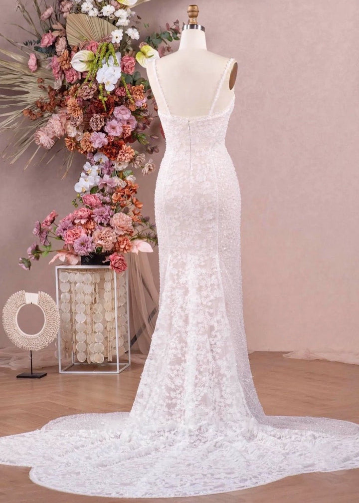 A fitted wedding dress with spaghetti straps, beading, lace and a flower cut-out train on a mannequin in front of a studio setting.