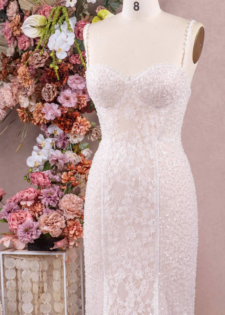 A close up of a fitted wedding dress with spaghetti straps, beading and lace on a mannequin in front of a studio setting.