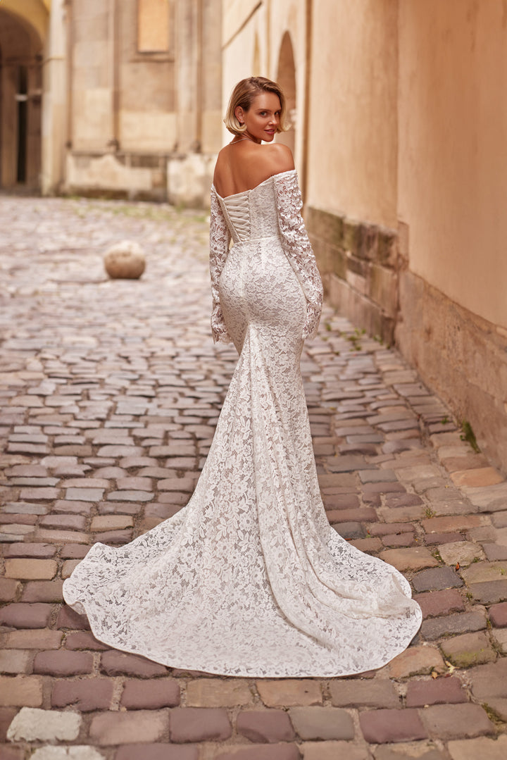 Woman in a fitted sleeved off-the-shoulder lace wedding dress with a lace up back and train standing against a rustic wall with plants.