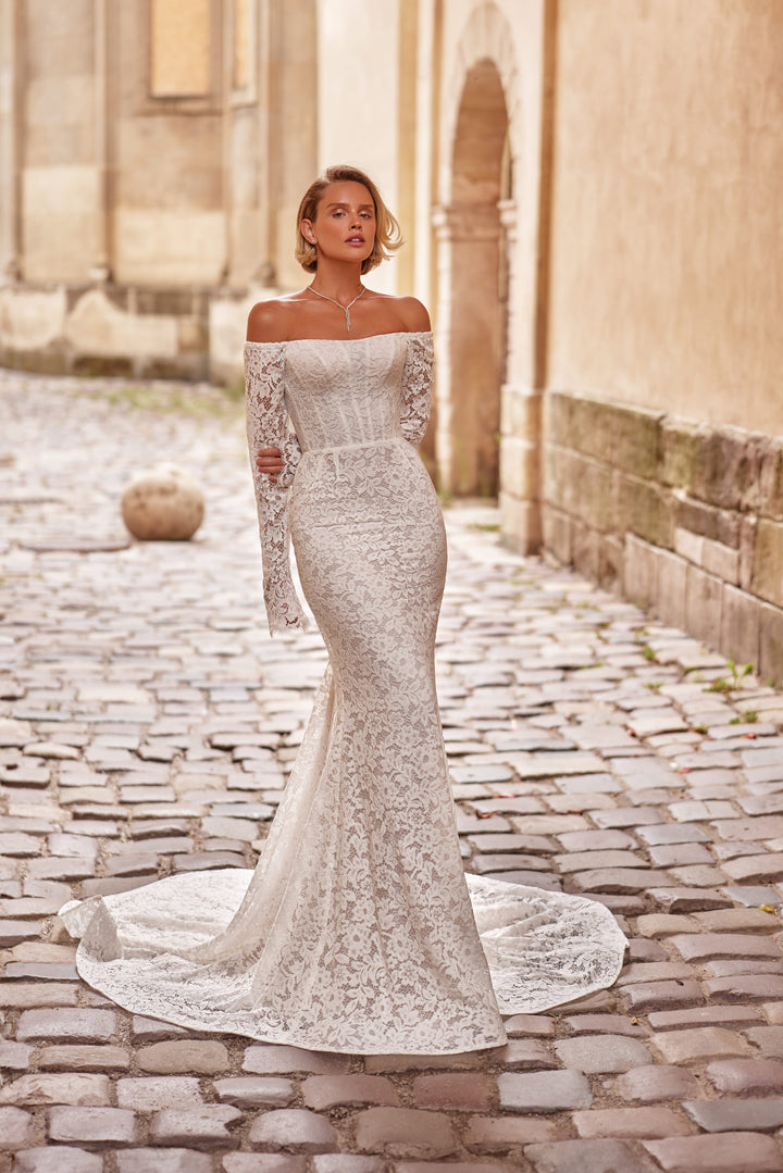 Woman in a fitted sleeved off-the-shoulder lace wedding dress standing against a rustic wall with plants.