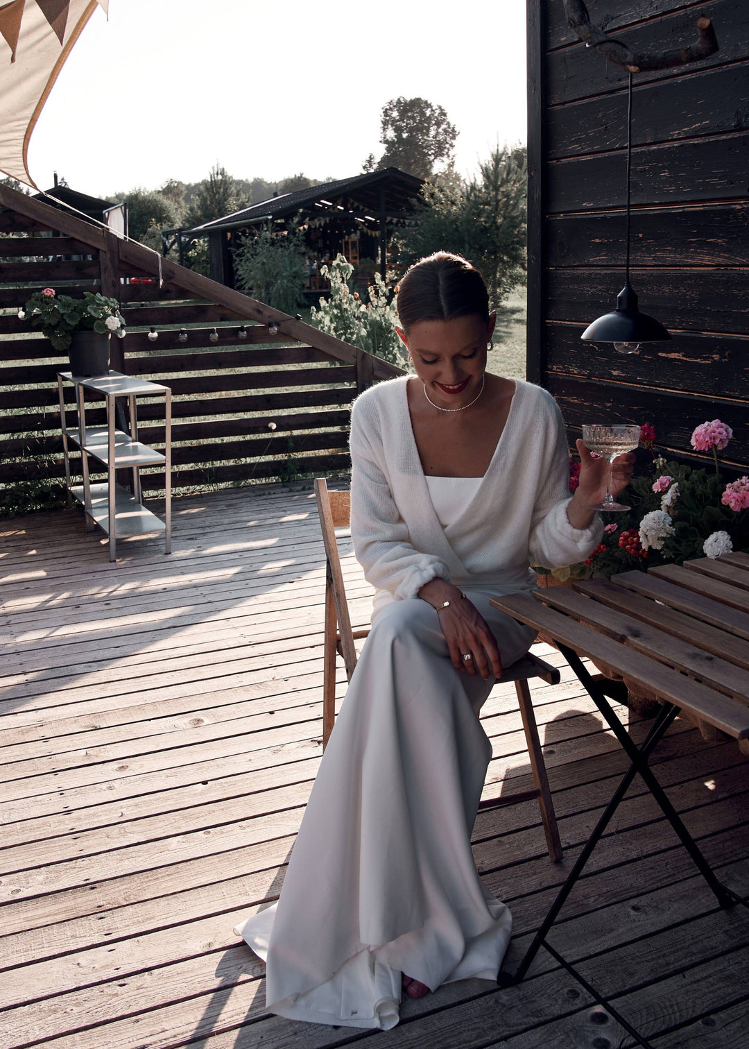 Woman in a white dress and warm wool bridal cardigan sitting on a wooden deck with a rustic background.