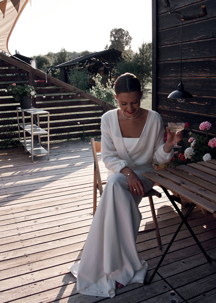 Woman in a white dress and warm wool bridal cardigan sitting on a wooden deck with a rustic background.