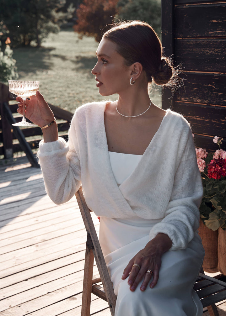 Woman in a white wedding dress and bridal cardigan holding a glass of champagne on a wooden deck.