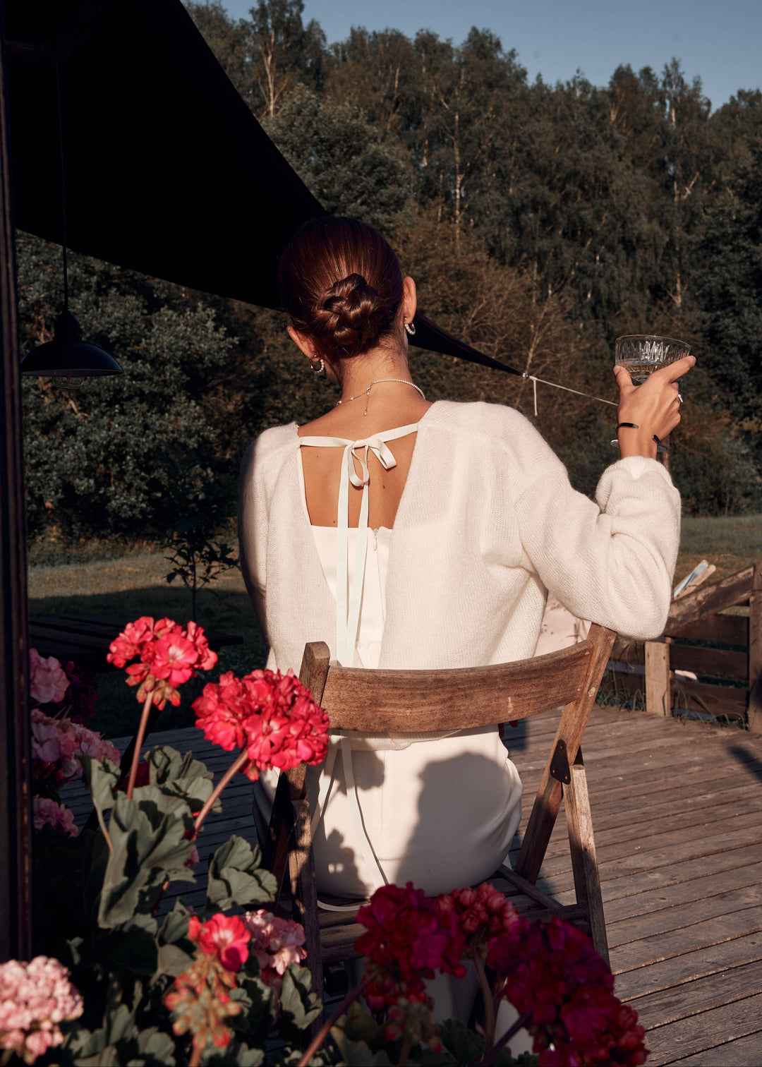 Woman in a white wedding dress with a tie up cardigan sitting on a chair with flowers and a scenic background.