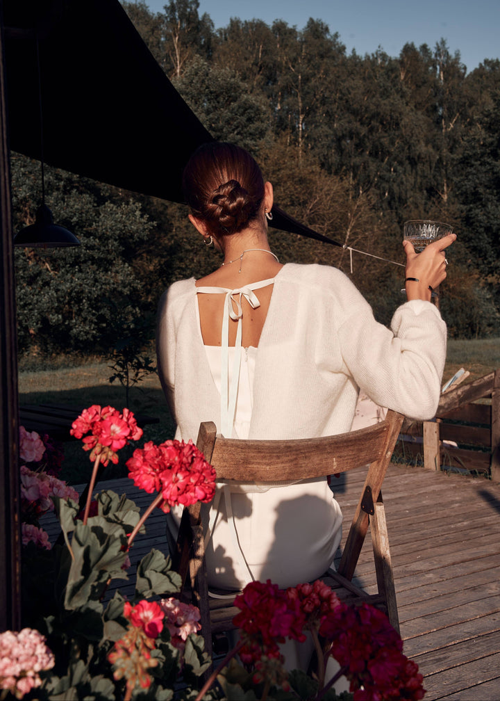 Woman in a white wedding dress with a tie up cardigan sitting on a chair with flowers and a scenic background.