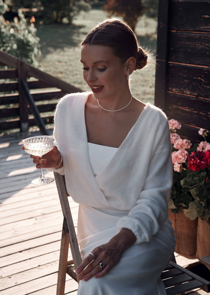 Woman in a white dress and warm bridal cardigan holding a glass on a wooden deck with flowers and trees in the background.