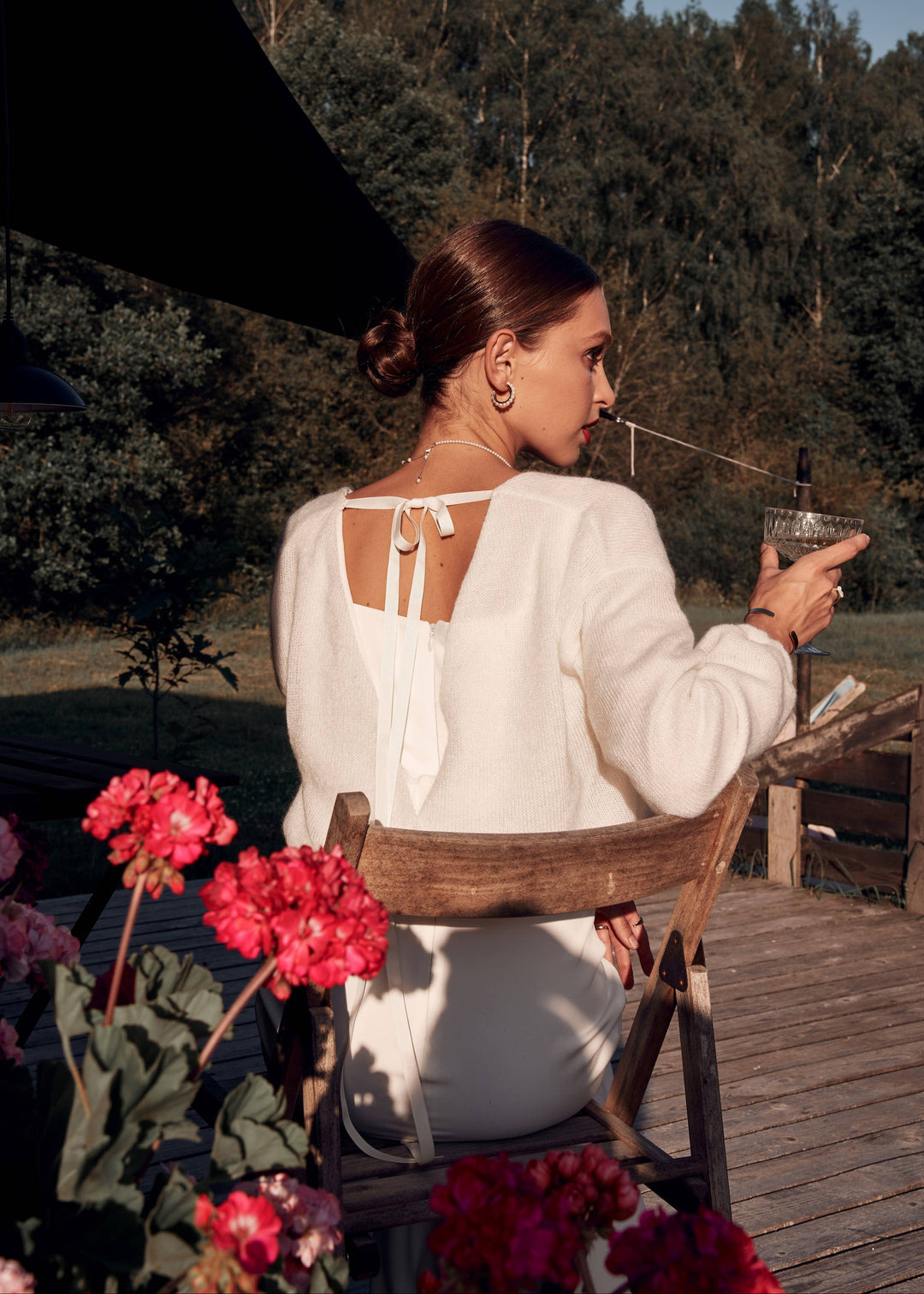 Woman in a white dress and warm wool cardigan sitting outdoors with flowers and a scenic background.