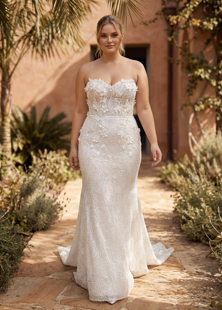 Woman in a strapless mermaid lace wedding gown standing with 3D flower appliques in a garden setting.