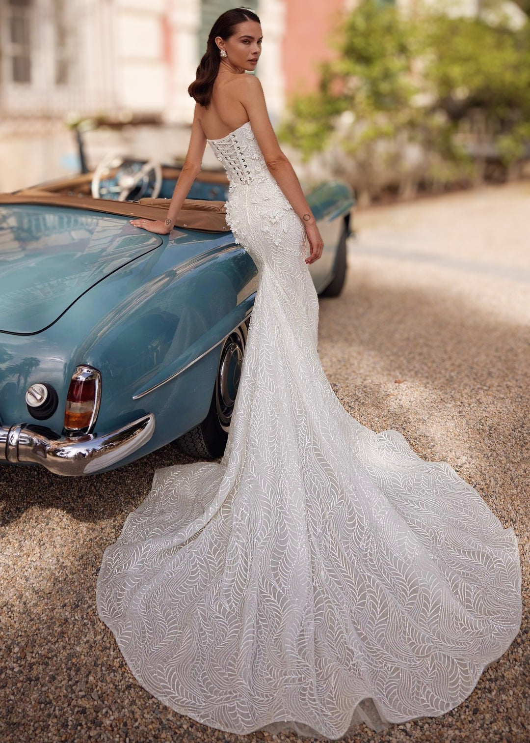 Woman in a strapless mermaid lace wedding gown with 3D flower appliques a lace up back and train standing leaning against a car.