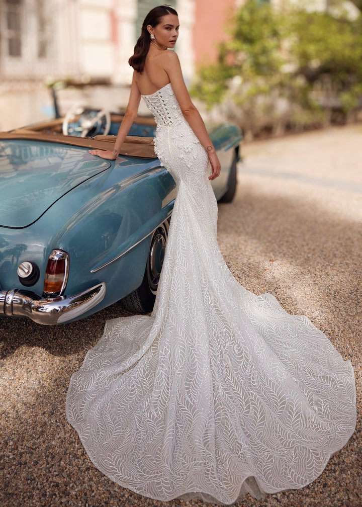 Woman in a strapless mermaid lace wedding gown with 3D flower appliques a lace up back and train standing leaning against a car.