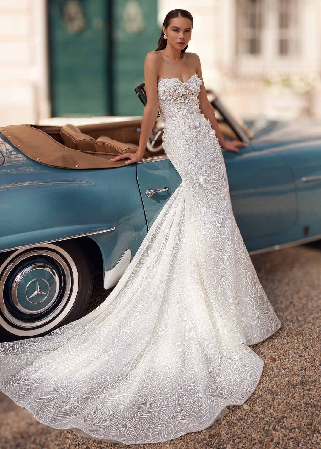 Woman in a strapless mermaid lace wedding gown standing with 3D flower appliques leaning against a car.