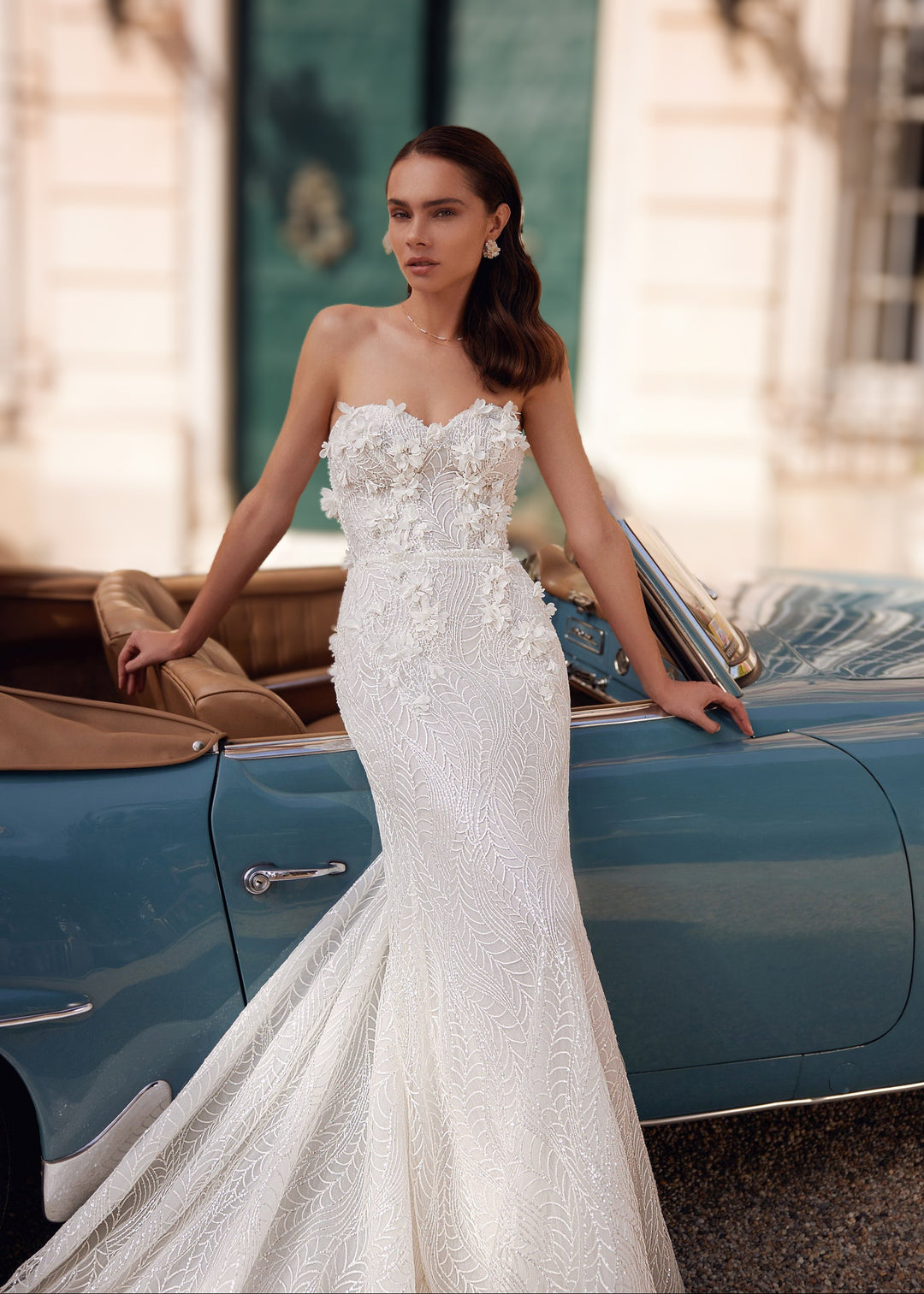 Woman in a strapless mermaid lace wedding gown with 3D flower appliques standing leaning against a car.