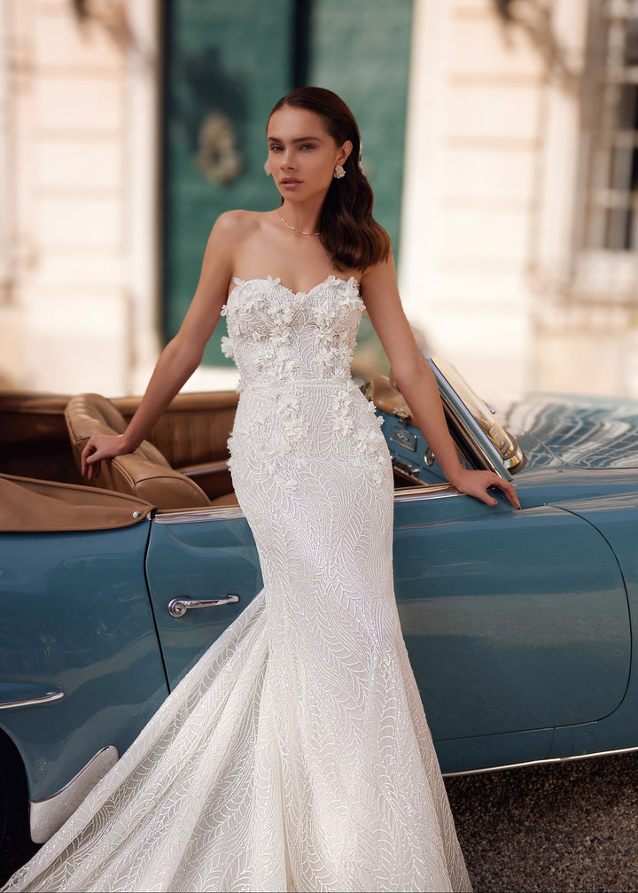Woman in a strapless mermaid lace wedding gown with 3D flower appliques standing leaning against a car.