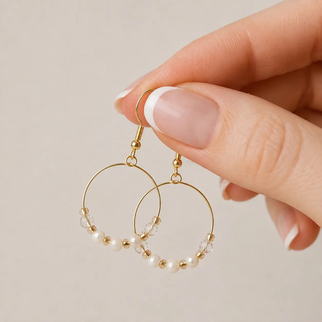Gold-plated hoop earrings with small pearls, clear bead accents and honey onyx beads, held by a hand with a French manicure against a soft beige background.