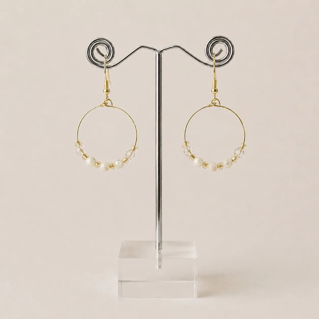 A pair of gold-plated hoop earrings with small pearls, honey onyx beads along the lower curve, displayed on a clear acrylic stand against a warm beige background.
