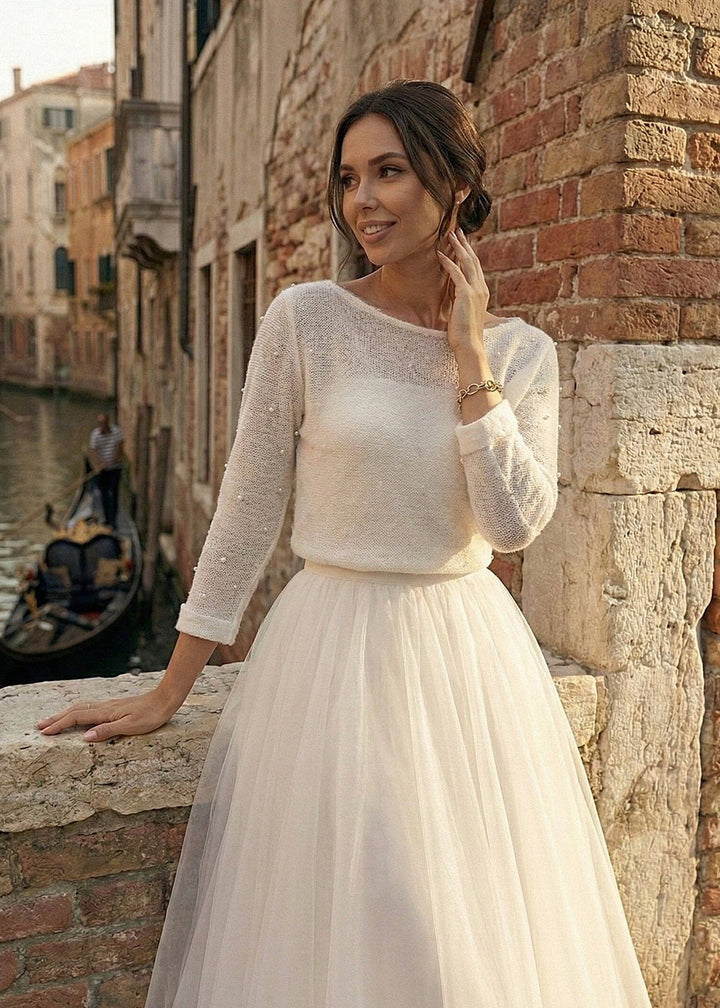 Woman in a white wedding dress wearing a knit cardigan, standing by a brick wall with a canal and gondola in the background