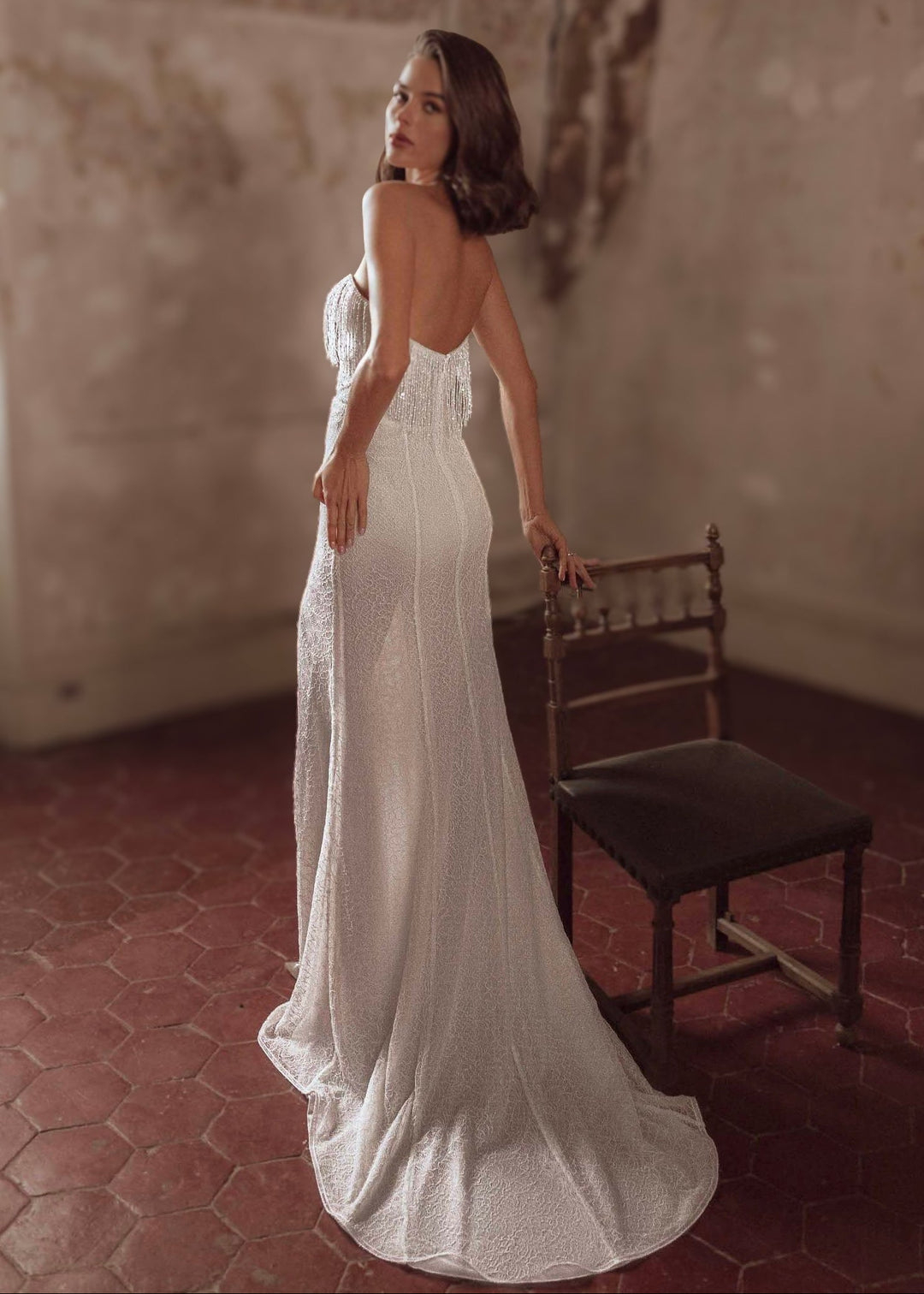 Woman wearing a strapless, lace and beaded wedding dress standing in a rustic room with a wooden chair.