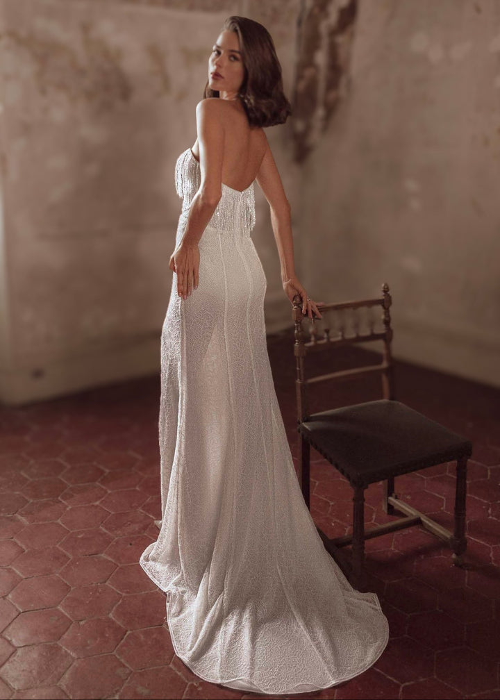 Woman wearing a strapless, lace and beaded wedding dress standing in a rustic room with a wooden chair.
