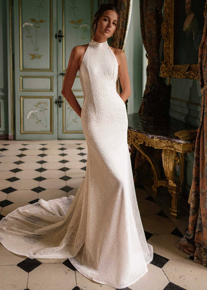 A woman standing in a room with antique decor, wearing a high neckline, sleeveless, white, guipure lace wedding dress with sequins and a sweeping train.