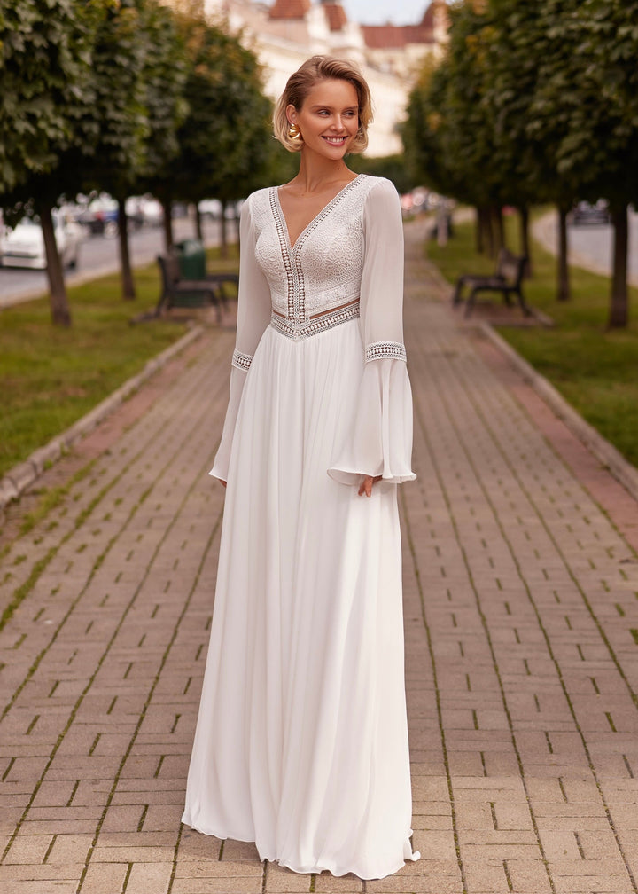 Woman in a boho chiffon wedding dress standing on a brick path with trees and benches in the background.