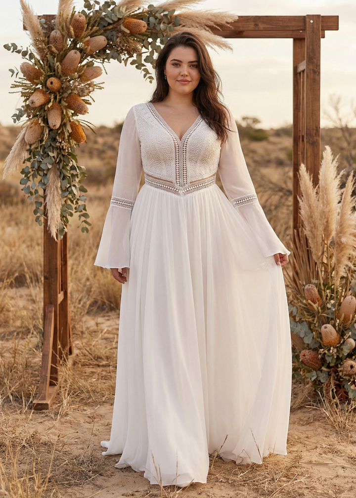 Woman in a white boho wedding dress standing in front of a decorative archway with flowers and pampas grass in a desert setting.