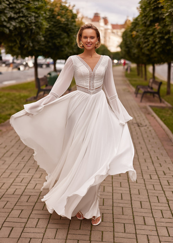 Woman in a white, long sleeve, a-line wedding dress standing on a brick path with trees and buildings in the background.