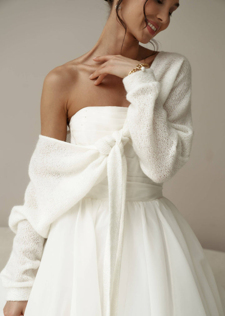Woman wearing a white wedding dress with a textured white cardigan against a neutral background.