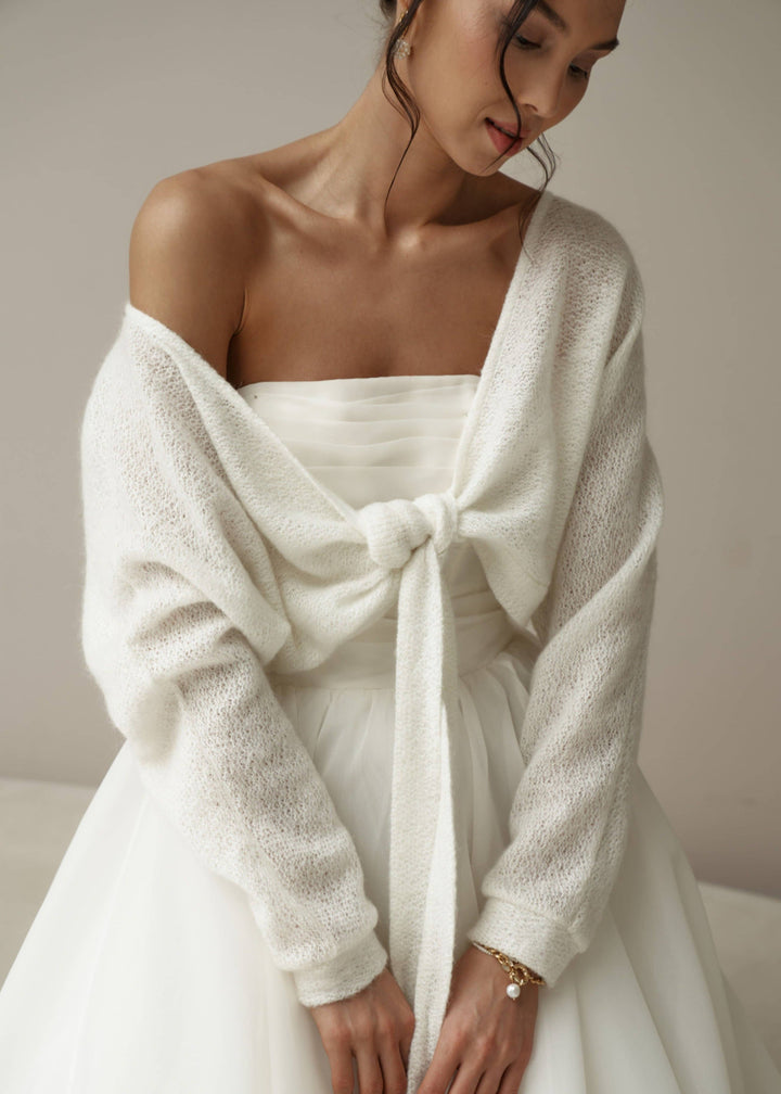 Woman wearing a white off-shoulder wedding dress with a mohair cardigan against a neutral background.