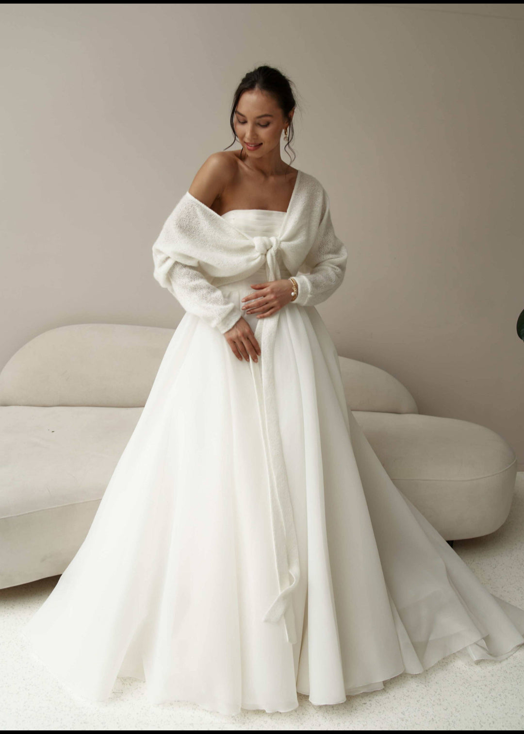 Woman wearing a white off-shoulder wedding gown with long sleeves cardigan in a minimalistic setting.