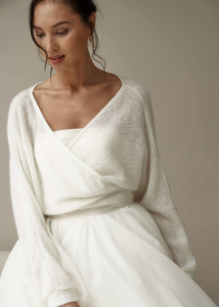 Woman wearing a white bridal dress with a multistyle cardigan against a neutral background.