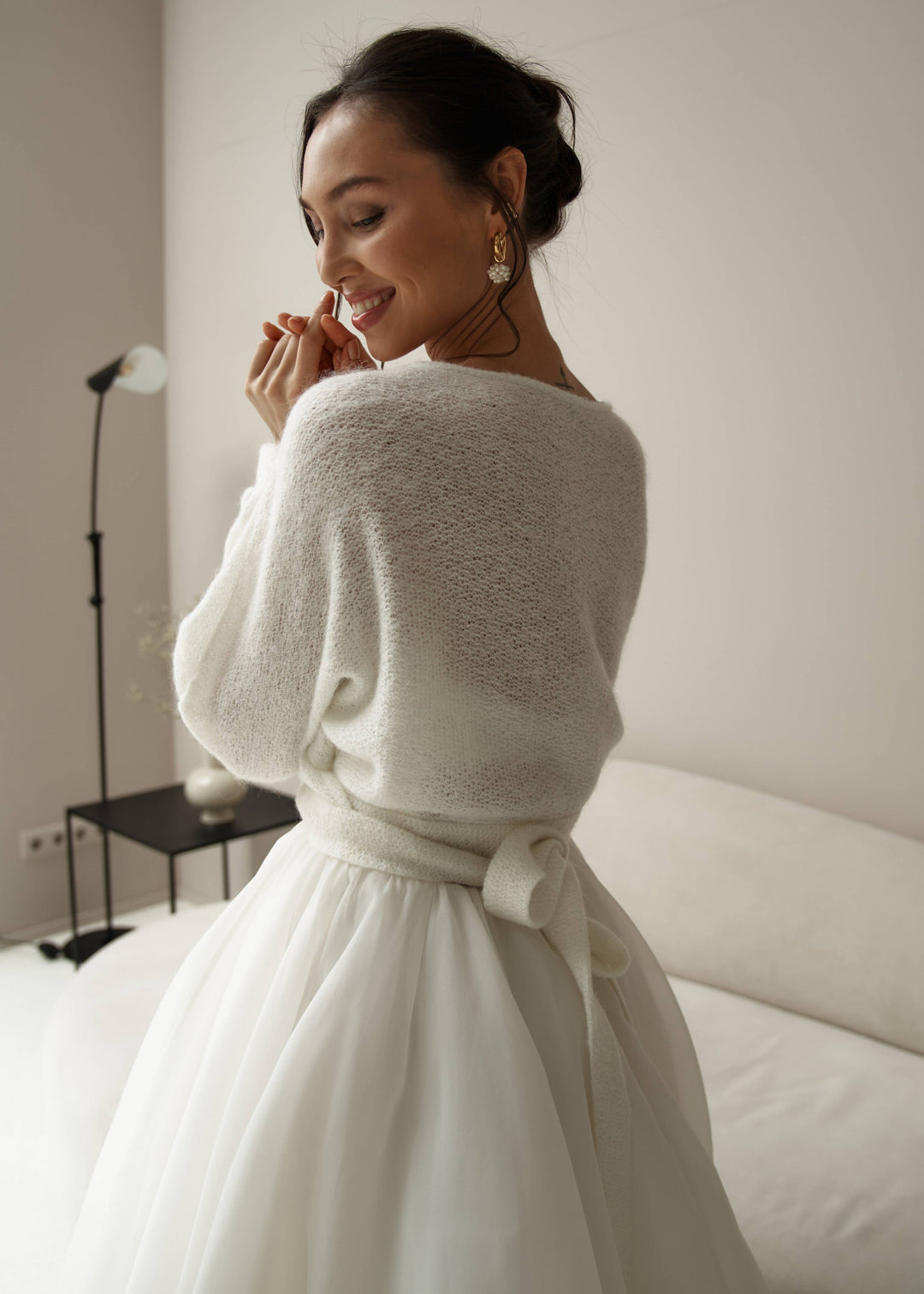 Woman wearing a white wedding dress with a warm mohair tie up cardigan against a plain background.