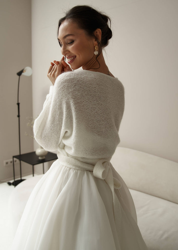 Woman wearing a white wedding dress with a warm mohair tie up cardigan against a plain background.