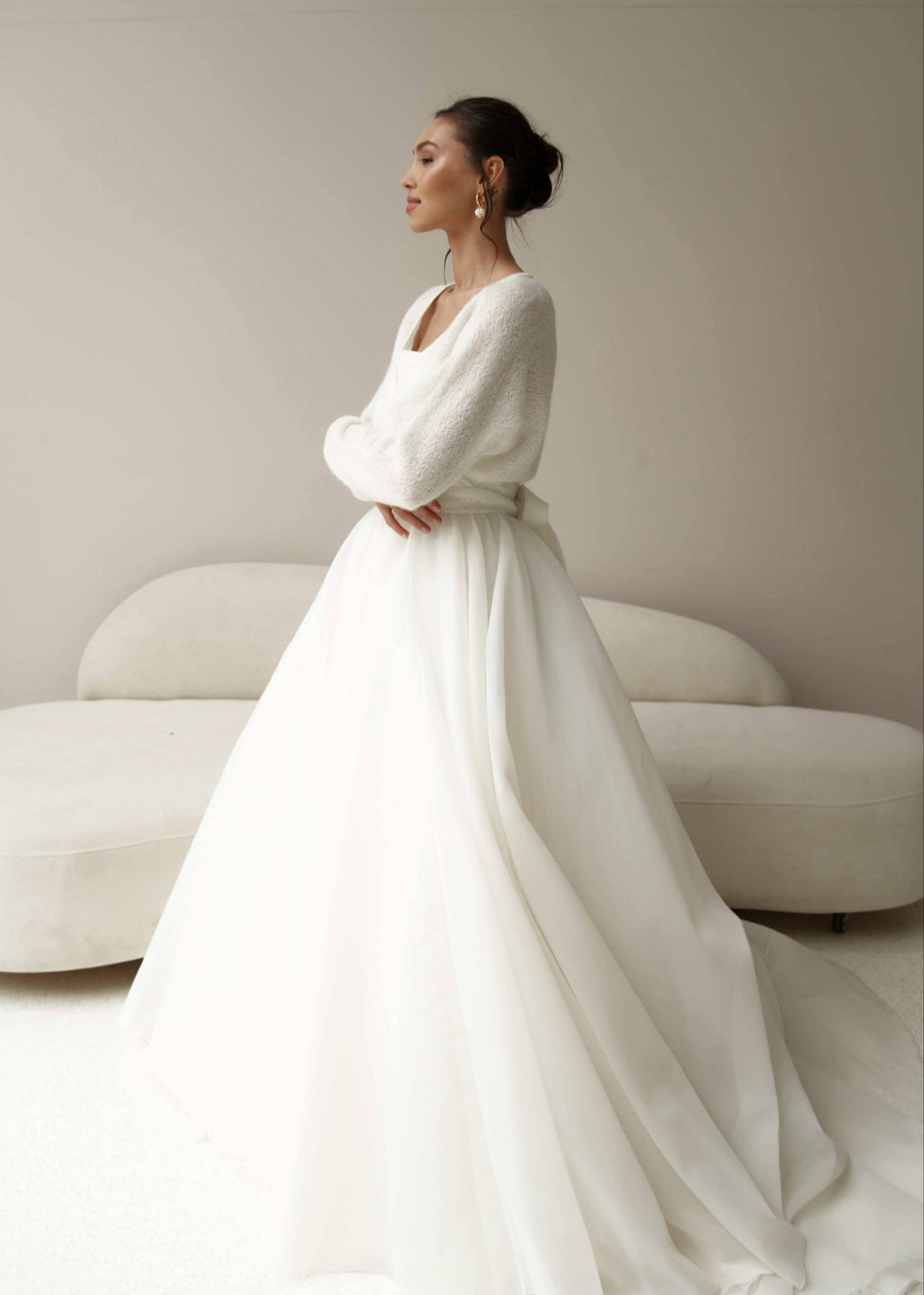 Woman in a white wedding dress and tie up ivory cardigan, standing against a neutral background.