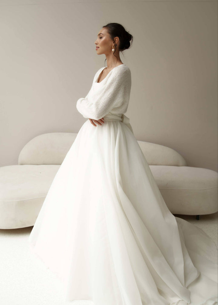 Woman in a white wedding dress and tie up ivory cardigan, standing against a neutral background.