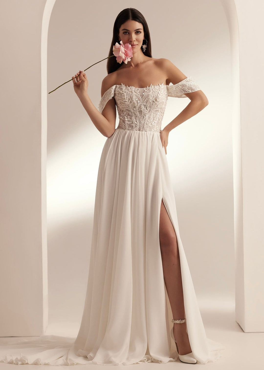 A woman in a white bridal gown with an off-shoulder neckline, 3D leaf appliqués, beads, sequins, and a front slit.