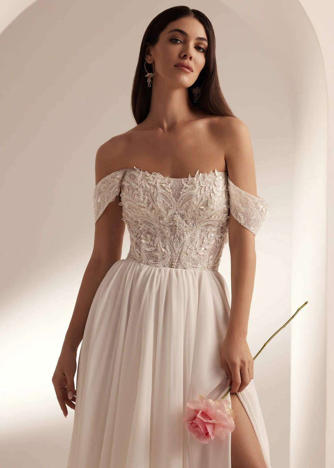 Woman wearing an off-the-shoulder wedding dress with a lace bodice and chiffon skirt against a plain background.