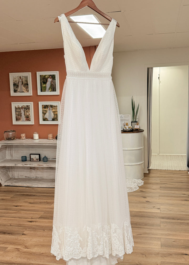White swiss-dot lace wedding dress hanging in a bridal store with wooden floor and framed pictures on the wall.